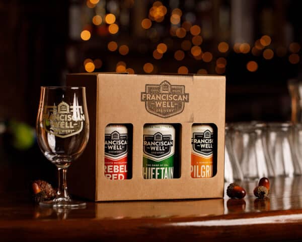 A Franciscan Well Bar beer gift box with three cans—Rebel Red, Chieftain, and Pilgrim—stands on a bar beside a branded glass, with warm lights and acorns in the background, capturing the spirit of their Products Archive.