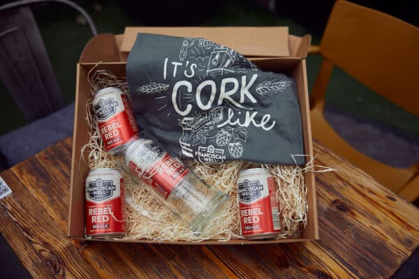 A gift box from the Products Archive featuring four cans of Franciscan Well Rebel Red beer, a drinking glass, and a dark t-shirt with It’s Cork like printed in white, all neatly arranged on straw in a cardboard box—perfect for any Franciscan Well Bar fan.
